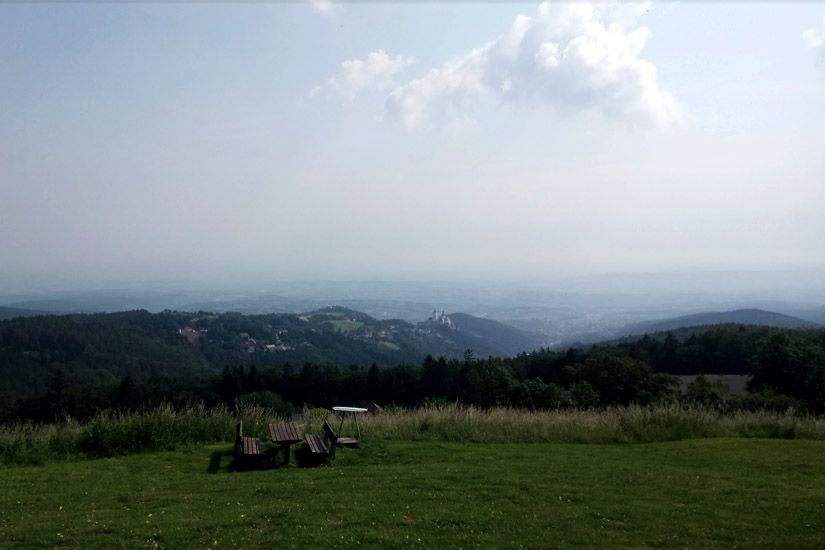 Magyarországi túra: Kilátás füves hegytetőről padokkal, erdő és távoli város felett, ideális hegyi túrázáshoz borús időben. Tekeregj.hu