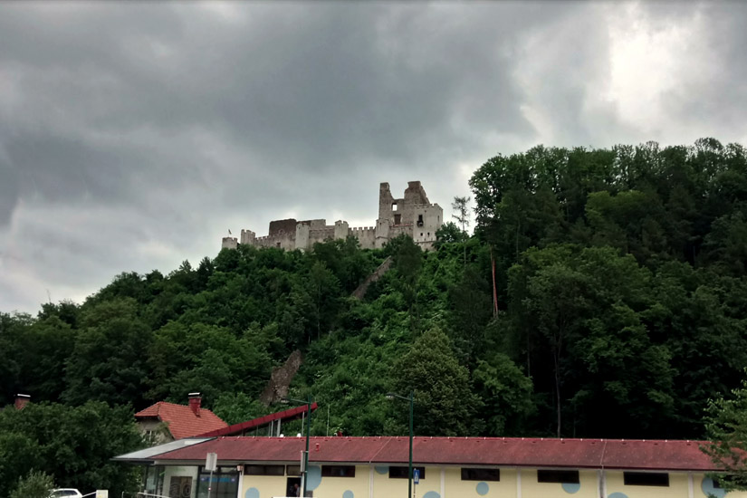 Magyarországi túra: Kővár rom a zöld, fás dombtetőn felhős ég alatt, túrázók kedvelt célpontja. Előtérben piros tetős, modern épület. Tekeregj.hu
