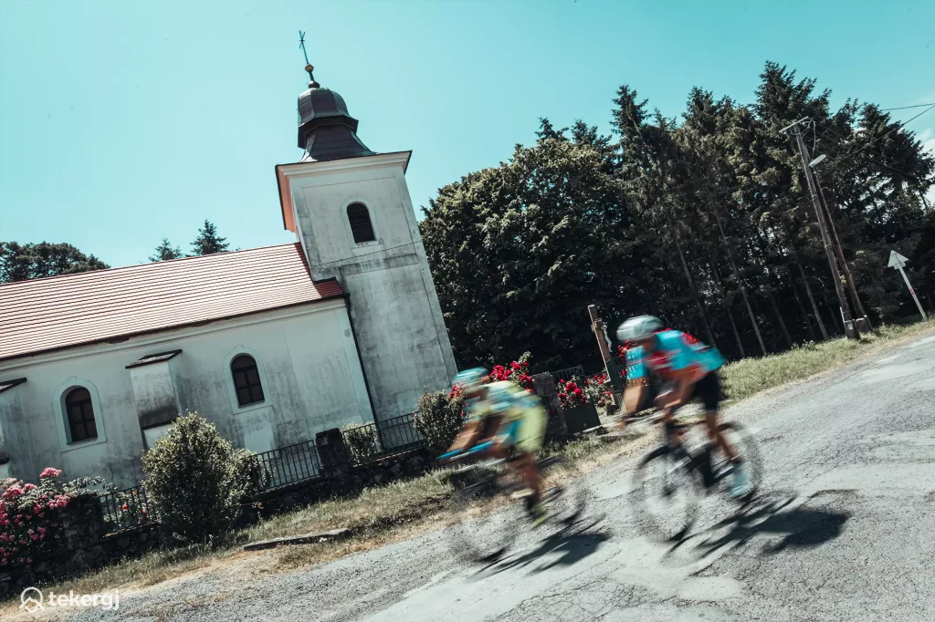 Magyarországi túra: Két hozás színes mezben teker egy fehér templom mellett napsütésben, természetjárás hangulatával, panorámás tájban. Tekeregj.hu