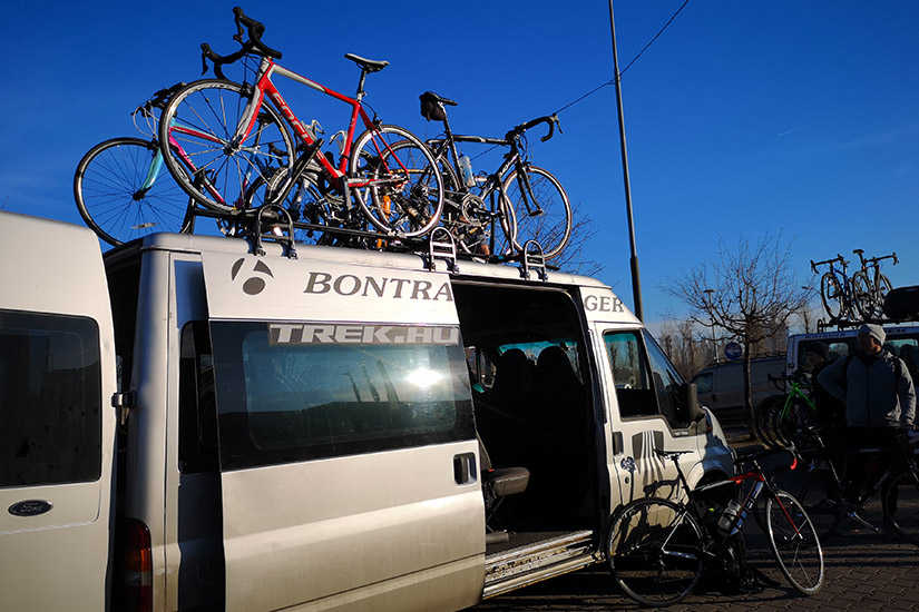 Magyarországi túra: Fehér furgon nyitott oldalsó ajtóval, tetején országúti bringákkal, hegyi túrára készülő csapat napos ég alatt. Tekeregj.hu