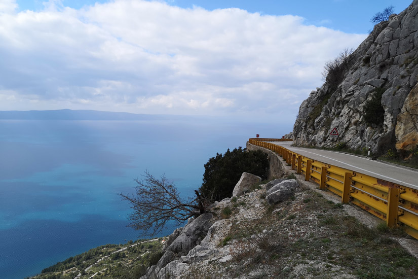 Magyarországi túra: Kanyargós, sárga korlátos tengerparti út sziklafal mellett, kilátással a kékre; ideális túrázáshoz vagy biciklizéshez. Tekeregj.hu