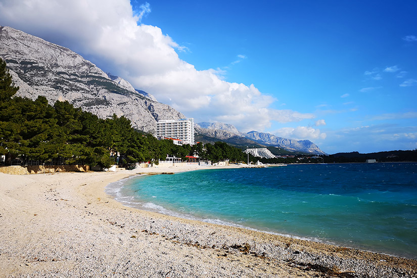 Magyarországi túra: Kavicsos strand türkiz vízzel, fenyvesekkel és hegyekkel, ideális természetjárásra; háttérben üdülők, kék ég. Tekeregj.hu