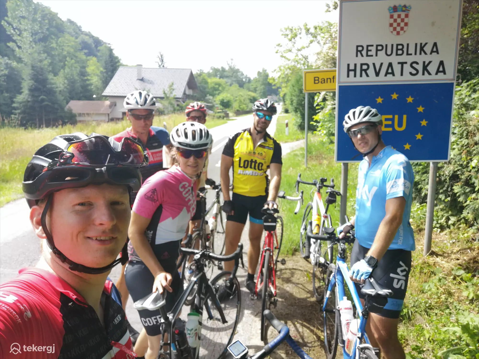 Magyarországi túra: Kerékpárosok pihennek a horvát-EU határjelző táblánál, napos időben, erdő és ház panorámájával. Tekeregj.hu