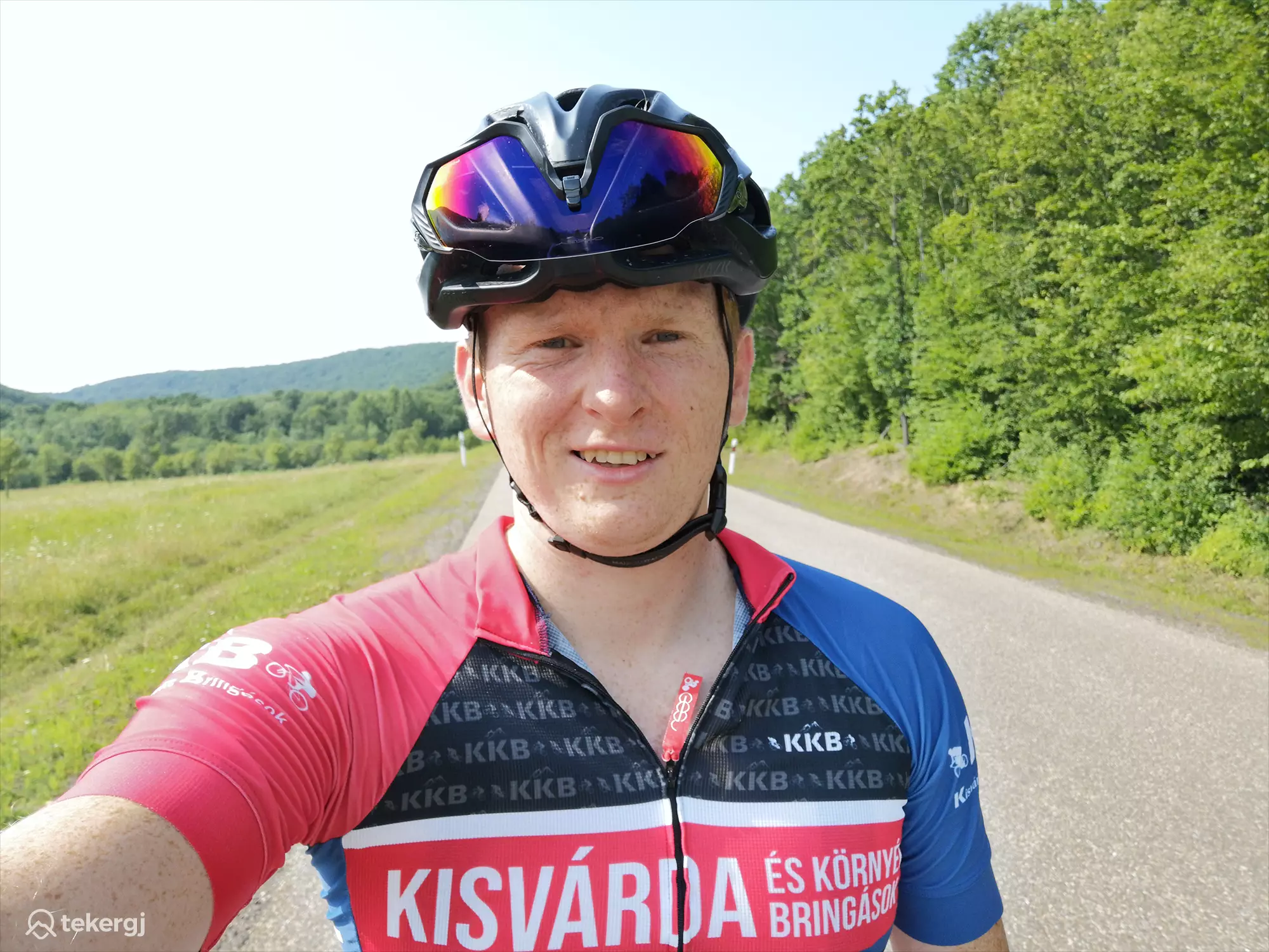 Magyarországi túra: Kerékpáros férfi sisakban, piros-kék mezben szelfizik hegyi túrán, zöld fák és dombok panorámájával, napsütésben. Tekeregj.hu
