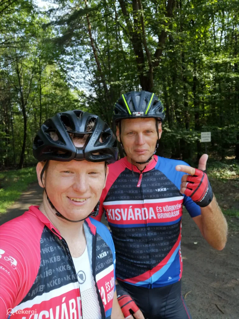 Magyarországi túra: Két kerékpáros piros mezben, bukósisakban szelfizik erdei ösvényen, napfényben. Tekeregj túra élmény. Tekeregj.hu
