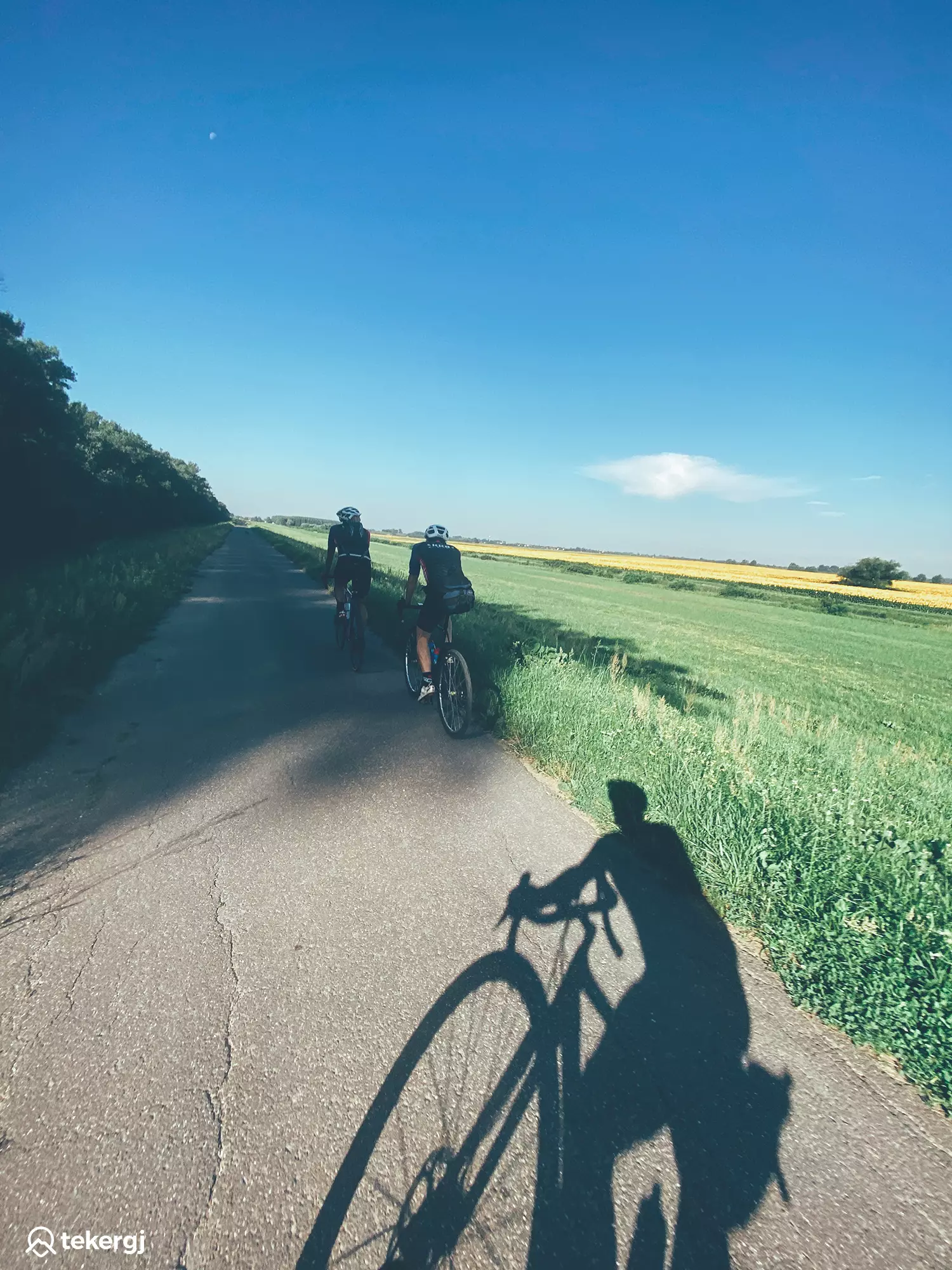 Magyarországi túra: Két kerékpáros teker zöld mezők között aszfaltúton, napos időben, panorámás tájjal és fák szegélyével. Tekeregj.hu