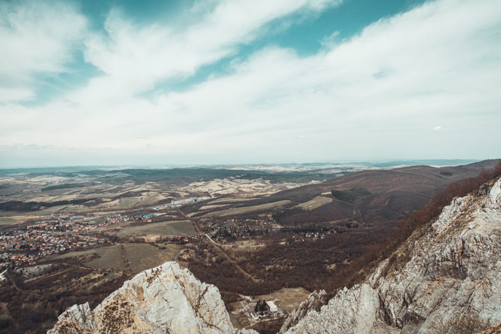 Magyarországi túra: Hegyi túrán készült panoráma: sziklás csúcs, völgy házakkal, dombok, erdők, kék ég részben felhőkkel. Tekeregj.hu