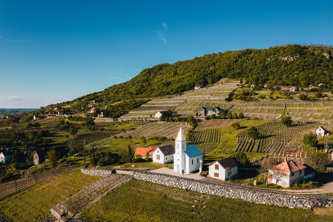 Magyarországi túra: Légi felvétel dombos vidékről, kis fehér templommal, szőlőkkel és házakkal; ideális hegyi túrához, természetjáráshoz. Tekeregj.hu