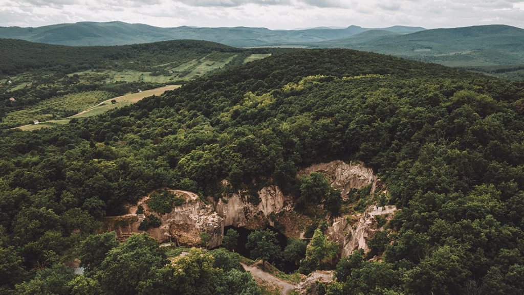 Magyarországi túra: Panoráma: zöld dombos erdő, középen nagy barlangbejárat, háttérben hegyek és felhős ég; ideális kirándulóhely. Tekeregj.hu
