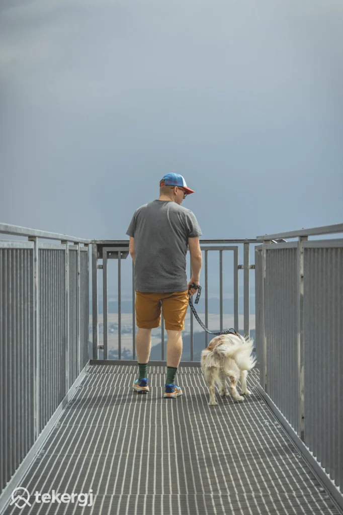 Magyarországi túra: Férfi sétáltat fehér kutyát fémsétányon, panorámás kilátással, felhős ég alatt. Bal alsó sarokban tekeregj logó. Tekeregj.hu