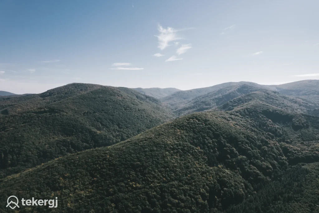 Magyarországi túra: Légi fotó zöld erdős dombokról tiszta kék ég alatt, távoli panorámával, tekeregj logóval a bal alsó sarokban. Tekeregj.hu