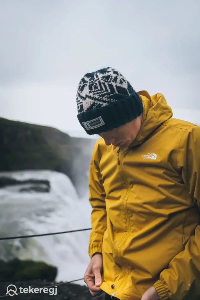 Magyarországi túra: Sárga North Face dzsekis túrázó esős időben vízesésnél, kirándulás közben, kezével a kabáton. Tekeregj.hu