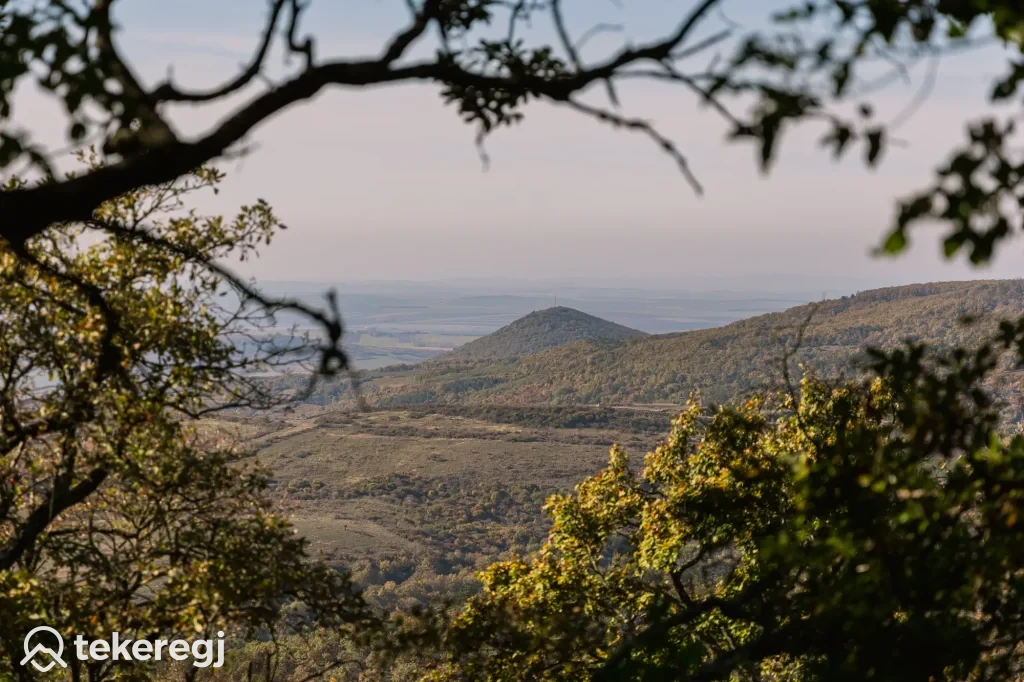 Magyarországi túra: Faágakon át látszik zöld domb és sík táj ködös ég alatt, ideális hely kiránduláshoz. Bal alsó sarokban Tekeregj logó. Tekeregj.hu
