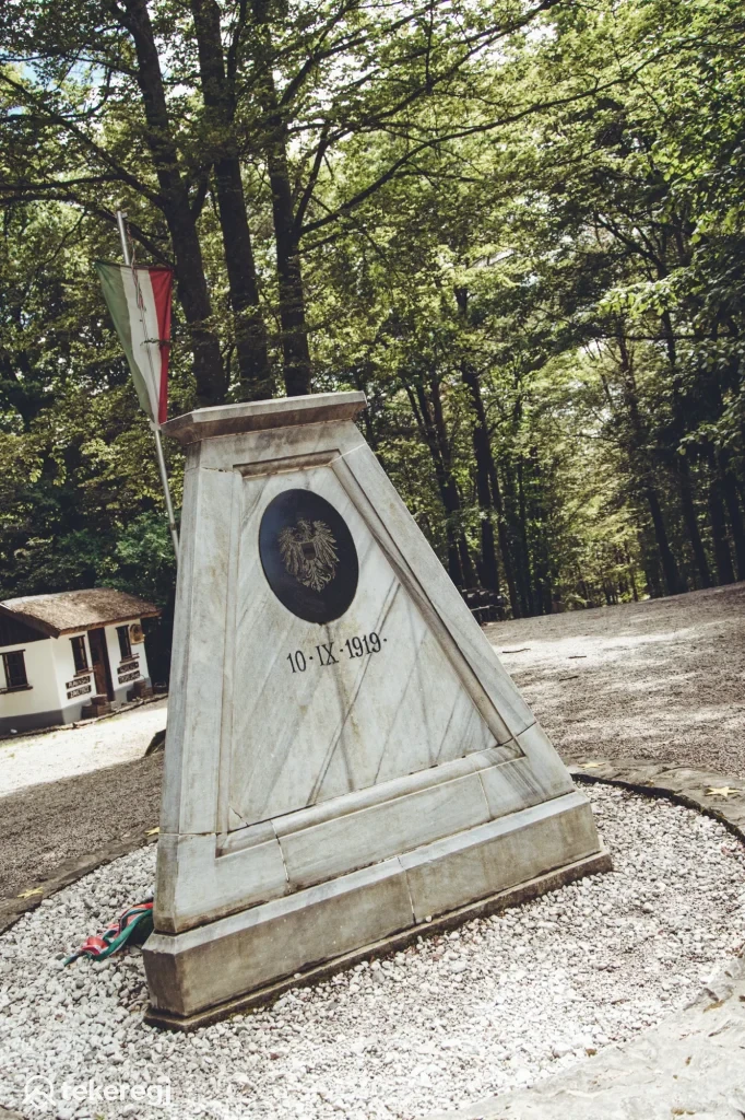 Magyarországi túra: Kő emlékmű sasdomborművel és olasz zászlóval erdei kirándulóhelyen, háttérben épület, panorámás kilátással. Tekeregj.hu