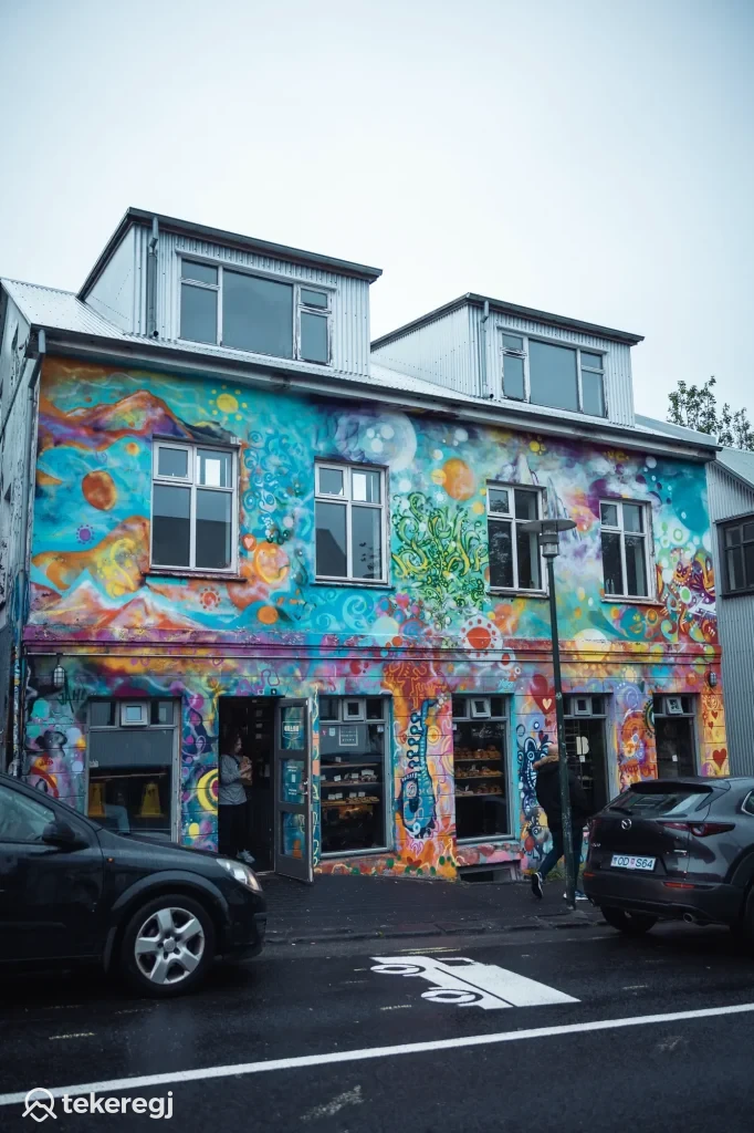 Magyarországi túra: Színes graffiti-s kétszintes ház előtt kiránduló sétál befelé, parkoló autók, felhős ég. Városi túraútvonal pihenő. Tekeregj.hu