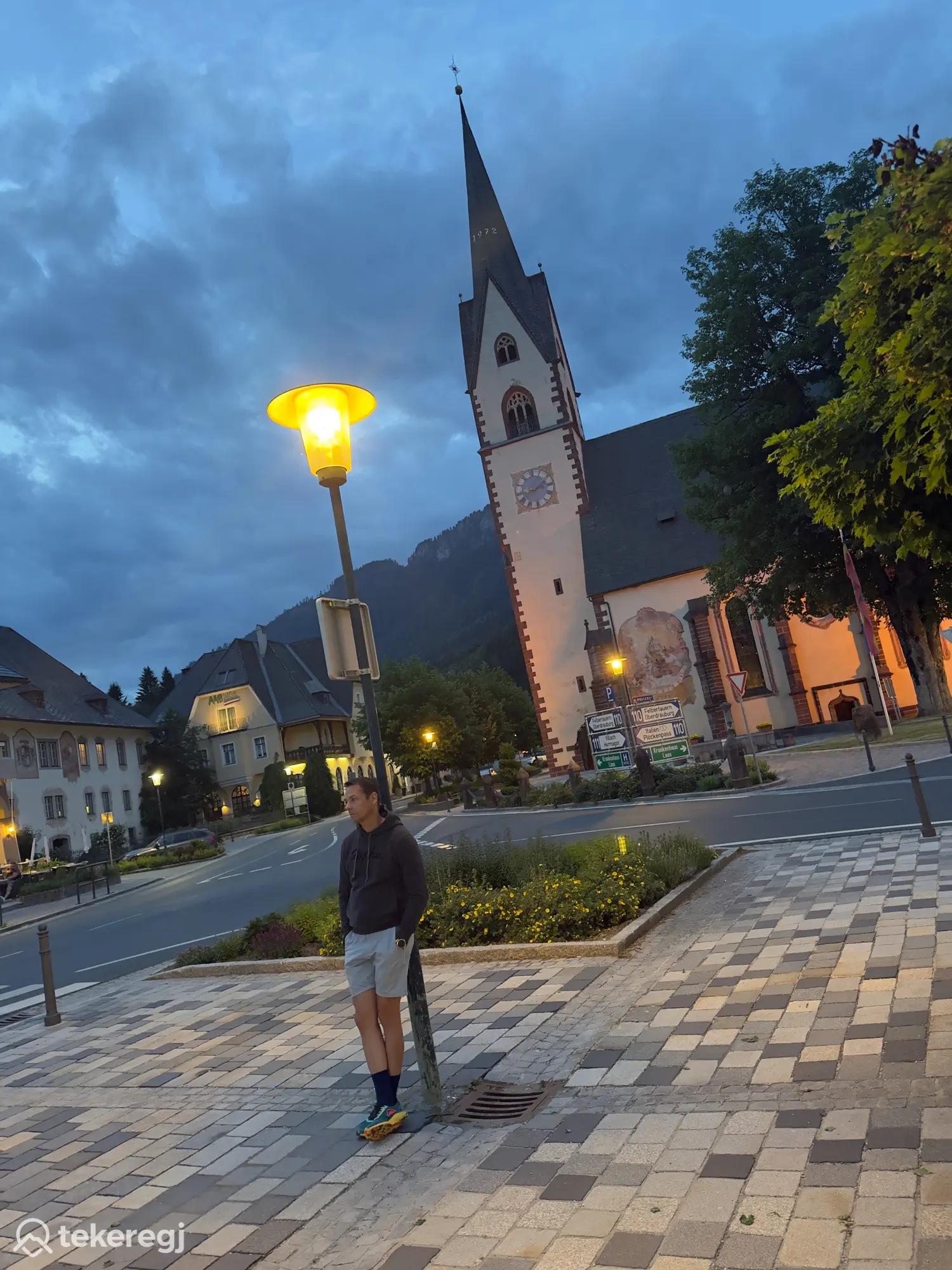 Magyarországi túra: Egy kerékpáros áll egy lámpa alatt egy osztrák kisváros utcáján alkonyatkor, a háttérben templomtorony és hegyek láthatók. Tekeregj.hu