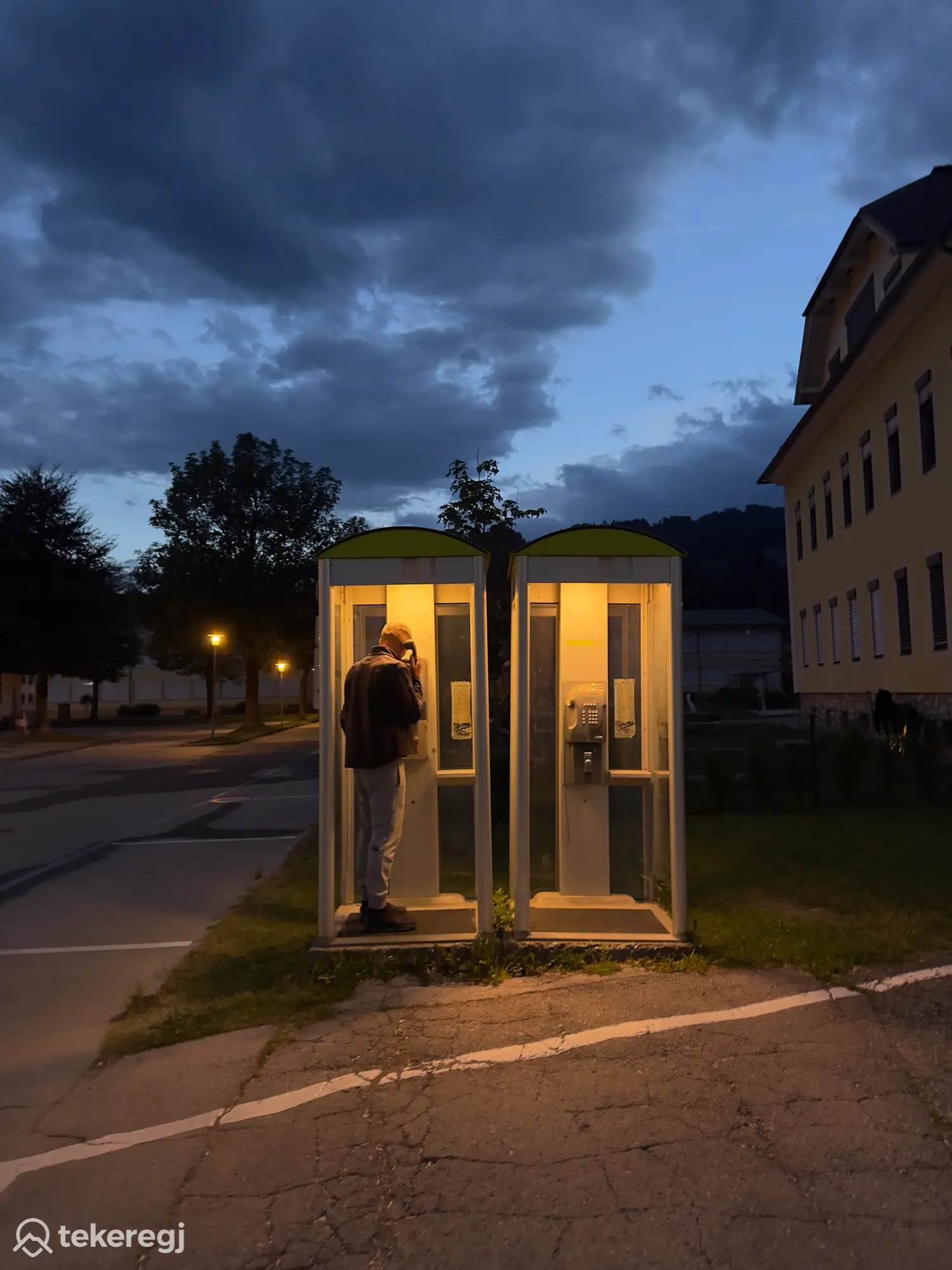 Magyarországi túra: Egy kerékpáros alkonyatkor megáll telefonálni egy osztrák telefonfülkében; mellette a kerékpárja áll, a csendes utcán fák és épületek látszanak. Tekeregj.hu