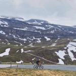 Magyarországi túra: Kerékpáros a Grossglockner-hágón, havas foltokkal és sziklás hegyoldalakkal, felhős ég alatt, az Alpok hágóinak felfedezése közben. Tekeregj.hu