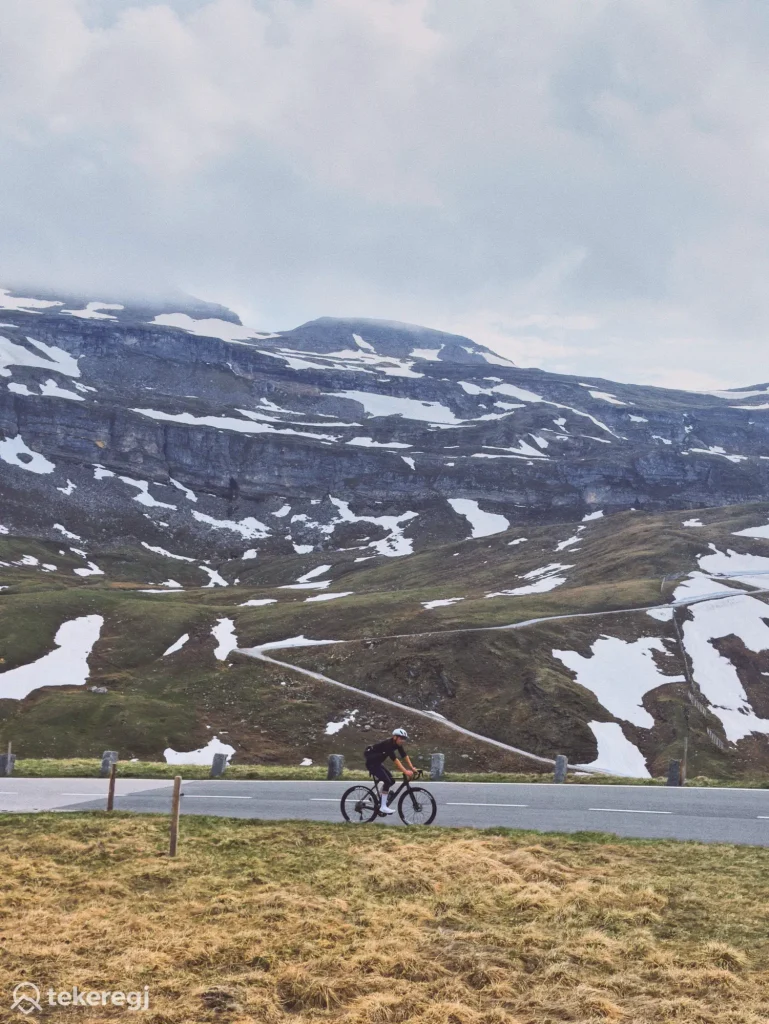Magyarországi túra: Kerékpáros a Grossglockner-hágón, havas foltokkal és sziklás hegyoldalakkal, felhős ég alatt, az Alpok hágóinak felfedezése közben. Tekeregj.hu