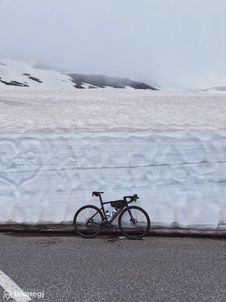 Magyarországi túra: Fekete kerékpár nedves úton, magas hófal előtt, a ködös Alpokban, a Grossglockner környékén zajló „Hágóvadászat” túrán. Tekeregj.hu