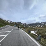 Magyarországi túra: Egy kerékpáros felfelé teker a kanyargós Grossglockner-i alpesi úton, zöld lejtők és havas foltok között, felhős ég alatt. Tekeregj.hu