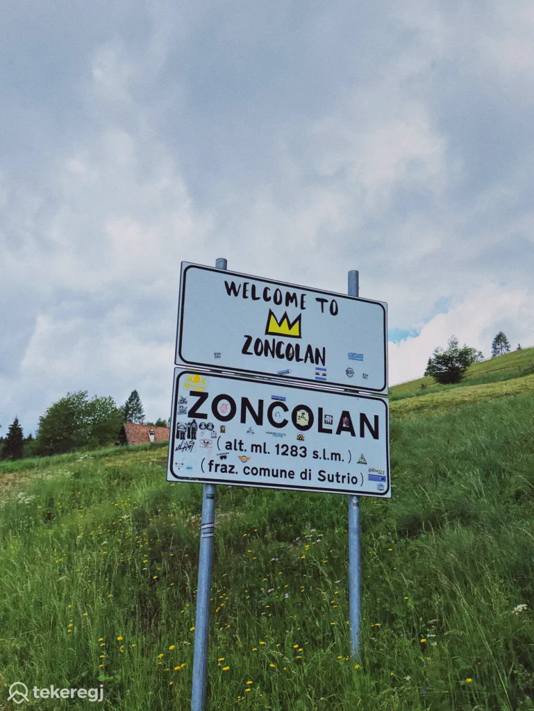 Magyarországi túra: A Monte Zoncolan táblája, matricákkal borítva, egy zöld domboldalon; magasság: 1283 m, kerékpárosok és túrázók kedvelt útvonalán. Tekeregj.hu
