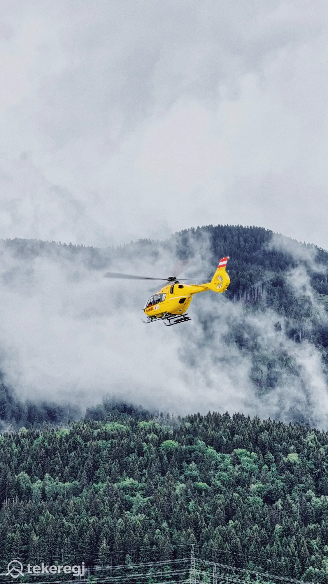Tekeregj túra: Egy sárga helikopter repül egy zöld erdő felett, háttérben ködös hegyek és felhős égbolt; túraútvonal az Alpokban. Tekeregj.hu